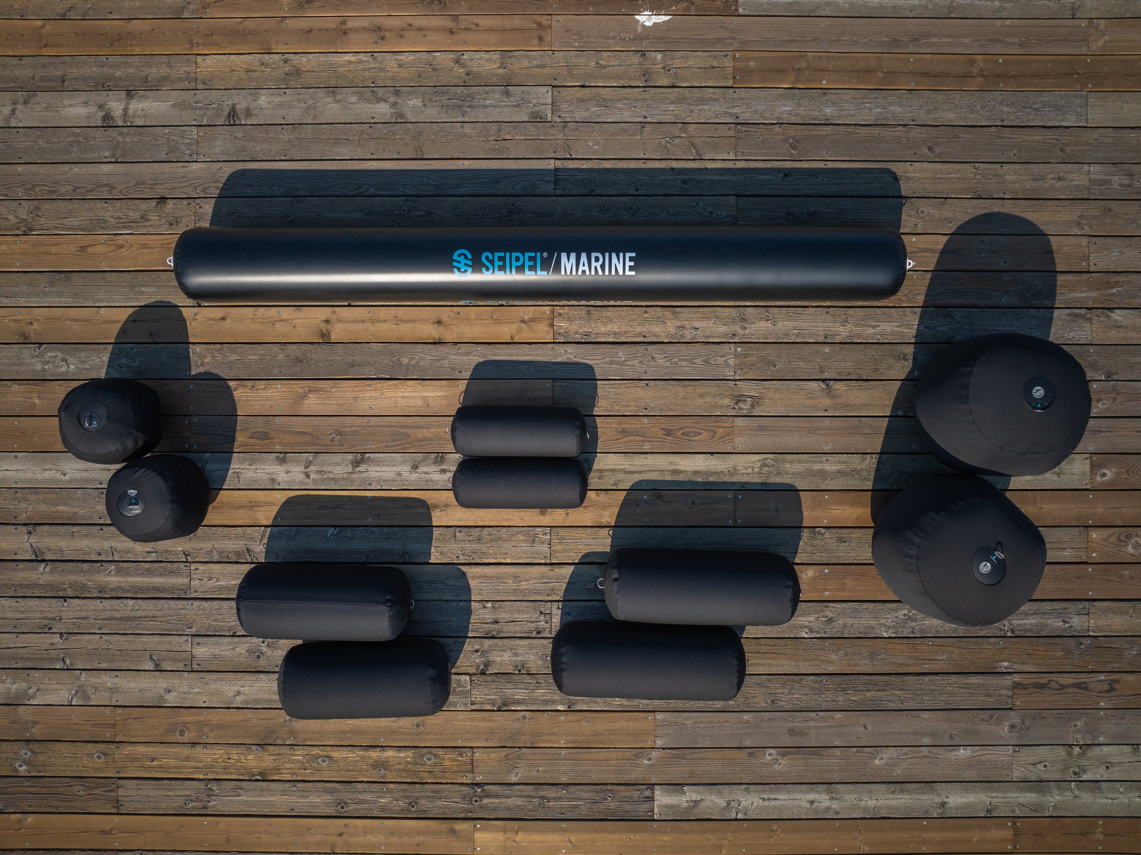 Top view of black Seipel Marine inflatable bumpers and fenders arranged on a wooden dock, with shadows cast by the sunlight.