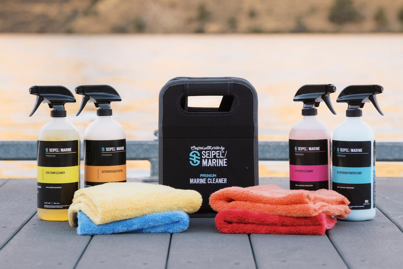 Seipel Marine cleaning kit with four spray bottles, a black carrying case, and microfiber towels on a dock at sunset.