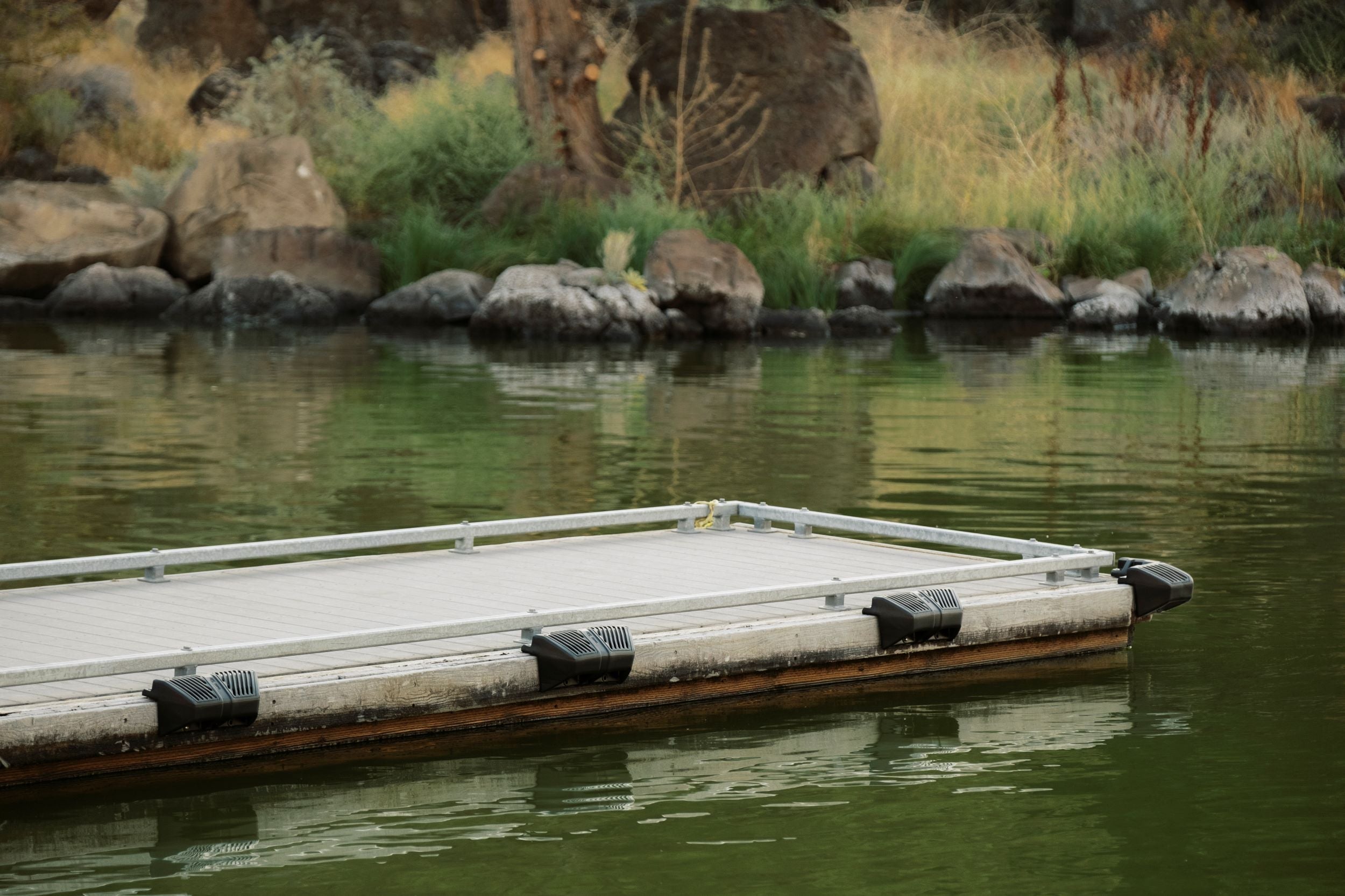 Close-up of dock with black corner bumpers on calm lake water near rocks and grass.