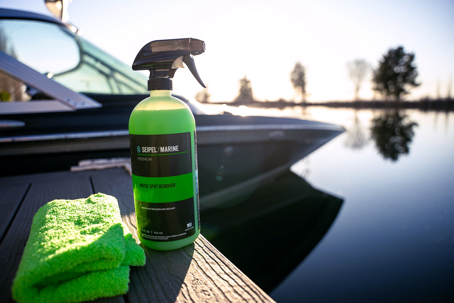 A bottle of Seipel Marine Water Spot Remover on a dock with a green microfiber cloth, with a boat and lake in the background.