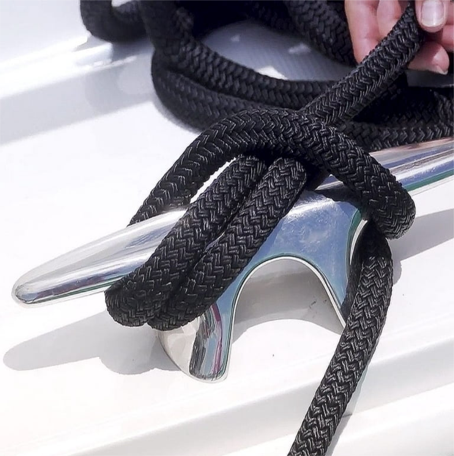 Close-up of a black braided dock line tied securely to a polished metal cleat on a white boat surface, with a hand adjusting the rope.