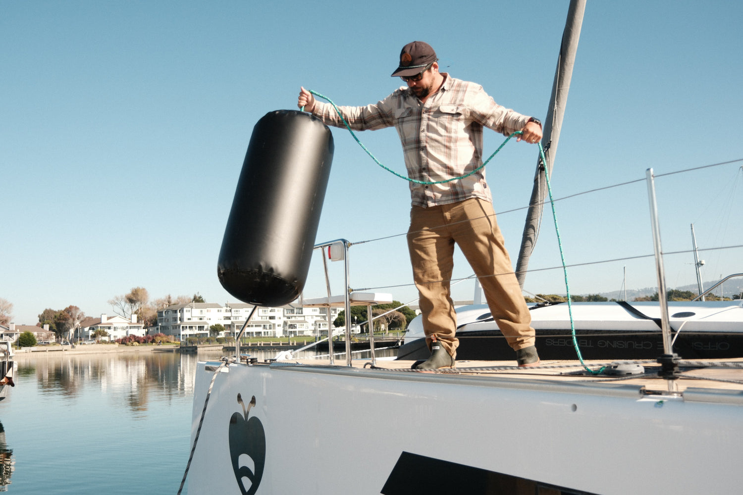 Person lifting a black inflatable fender onto the side of a catamaran at a marina.